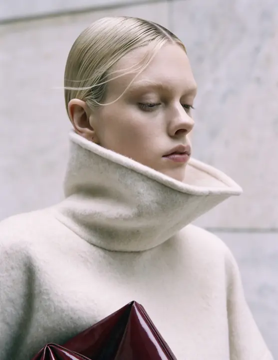 Ivory funnel-neck coat with a burgundy clutch against marble
